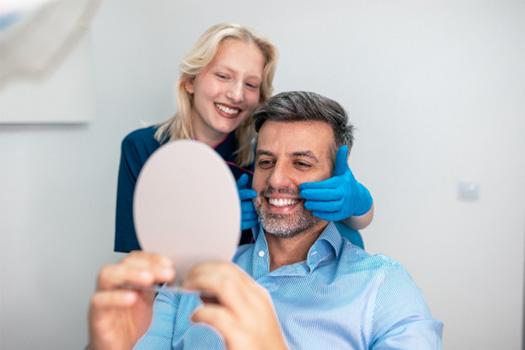 Man smiling at his reflection as dentist touches the corners of his mouth