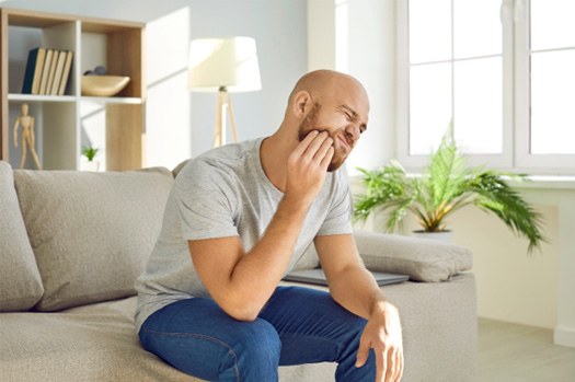 Man sitting on sofa squinting and pressing hand to jaw in pain